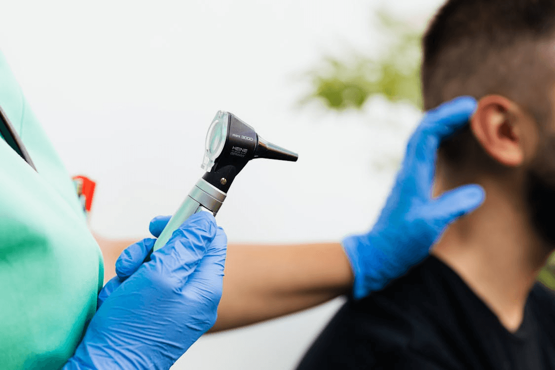a person wearing green clothes and medical gloves holding a Heine otoscope