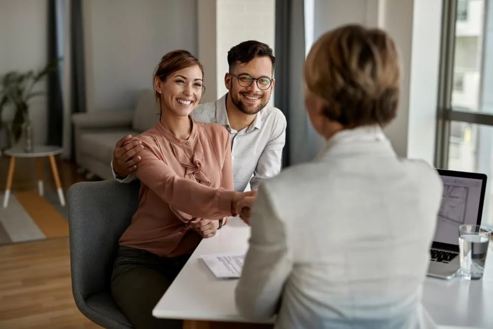 A smiling couple shaking hands with a professional health insurance broker across a desk during a successful consultation.